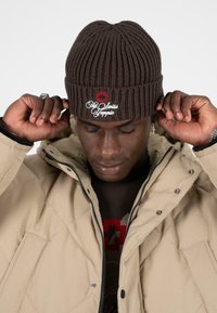 Bruine gebreide beanie met een geribbelde textuur, voorzien van een geborduurd rood kruis en de tekst "The Swiss Hippy". Draagt bij een beige gewatteerde jas.