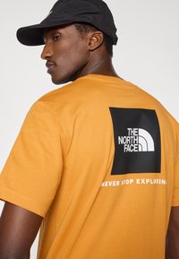 Μουσταρδί κίτρινο μπλουζάκι με μαύρο τετράγωνο γραφικό που αναγράφει "THE NORTH FACE" και το σύνθημα "NEVER STOP EXPLORING" σε λευκό. Ύφασμα από βαμβακερό μείγμα.