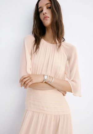 Mujer con cabello largo y castaño lleva un vestido transparente de color rosa pálido con mangas largas, pulseras plateadas en la muñeca, de pie frente a un fondo blanco.