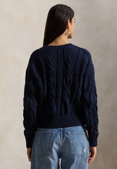 Μπλε πλεκτό πουλόβερ με σχέδιο πλεξούδας, ριμπ τελείωμα και μακριά μανίκια. Φορεμένο με ανοιχτόχρωμα τζιν παντελόνια με πίσω τσέπες.