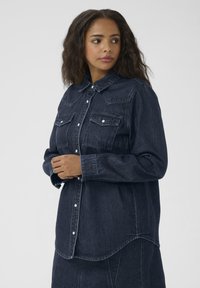 Dark denim shirt with a button-down front, two chest pockets, long sleeves, and a curved hem. Features a classic collar and contrast stitching.