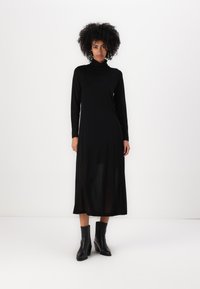Crna haljina s dugim rukavima i izrezom na turtleneck od laganog materijala, opuštenog kroja koji pada do sredine lista, u kombinaciji s crnim gležnjačama.