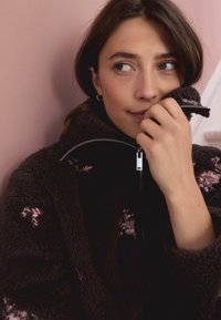 Juoda pūkuota džemperinė striukė su gėlėmis siuvinėjimu. Minkšta tekstūra, aukšta apykaklė ir sidabrinis užtrauktukas. Modelis su nuleistais plaukais laiko apykaklę.