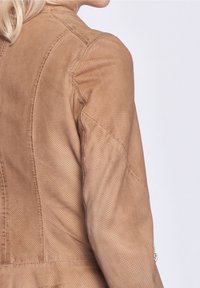 Hellbraune Lederjacke mit perforierter Textur, die ein tailliertes Design aufweist und sichtbare Nähte entlang der Schulter- und Rückennähte hat.