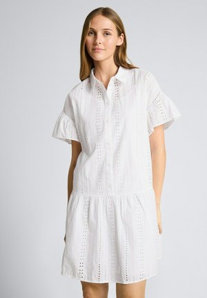 Jeune femme portant une robe blanche à manches courtes avec œillets, boutons et ourlet volanté, debout devant un fond clair uni.