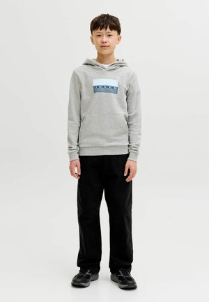 Zēns stāv ar seju pret priekšu, valkājot pelēku kapuci ar "JONES" logotipu, melnas bikses un melnas sporta kurpes uz vienkārša gaiša fona.