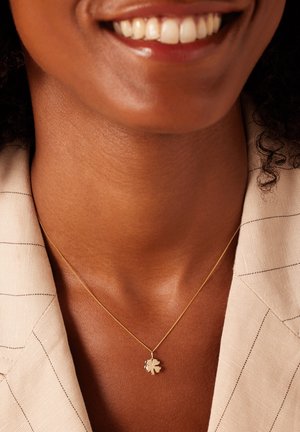 Collier en or avec un petit pendentif en forme de trèfle à quatre feuilles, porté sur une peau nue, sur un fond de tissu à rayures claires.