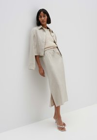 Beige Leinenjacke mit Kragen, kombiniert mit einem gestrickten cremefarbenen Oberteil und einem passenden Bleistiftrock mit seitlichem Schlitz, ergänzt durch weiße Sandalen.