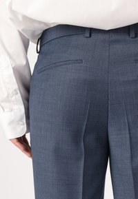 Blaue gemusterte Hose aus strukturiertem Stoff, mit flachem Bund, zwei Gesäßtaschen und tailliertem Schnitt, kombiniert mit einem weißen Hemd.