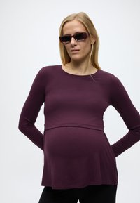 Έγκυος γυναίκα με μακριά ξανθά μαλλιά, φορώντας μακρυμάνικη μπλούζα σκούρου μοβ και γυαλιά ηλίου στο ίδιο χρώμα, στέκεται με τα χέρια πίσω από την πλάτη.