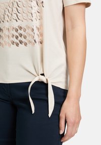 Lyse beige kortærmet t-shirt med knude detalje i kanten og et reflekterende geometrisk mønster på forsiden. Blød stofstruktur.