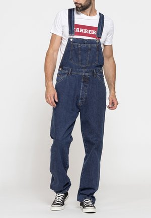 Uomo con salopette in denim blu navy, maglietta bianca con scritte rosse e scarpe da ginnastica nere, in piedi davanti a uno sfondo chiaro uniforme.