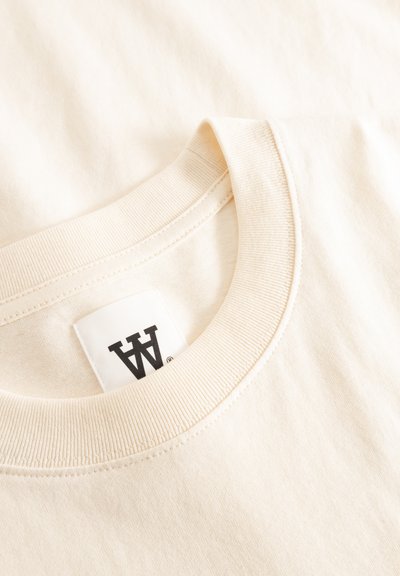 Μπεζ βαμβακερό T-shirt με ριμπ λαιμόκοψη, που έχει μια μαύρη ετικέτα λογότυπου σε λείο ύφασμα, διπλωμένο ώστε να αποκαλύπτει υφή και λεπτομέρειες.