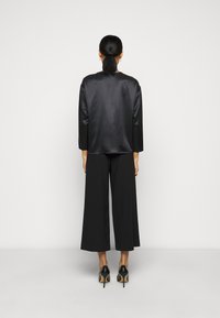 Veste en satin noir à manches longues, associée à un pantalon noir taille haute à jambes larges. Le modèle porte des talons hauts noirs, vue de dos affichée.