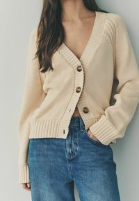 Frau in einer beigefarbenen, gestrickten Knopfpullover mit großen Knöpfen, kombiniert mit blauen Jeans, Hand in der Tasche, vor einem schlichten Hintergrund.