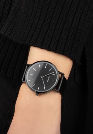 Black wristwatch with a smooth round face, white hour markers, and a black leather strap. The background features a textured black fabric.
