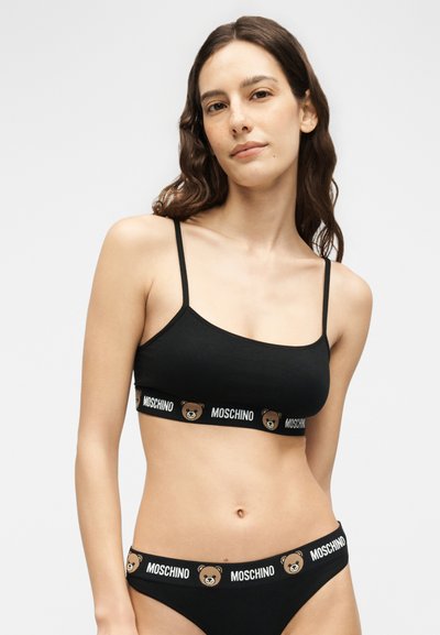 Νέα γυναίκα που φοράει μαύρο σετ σουτιέν και εσώρουχο Moschino με λογότυπα αρκουδάκια στα ελαστικά λουριά, στέκεται μπροστά σε απλό φόντο.