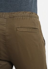 Olivgrön byxor med en slät yta, elastisk midja och en bakficka med ett horisontellt öppnande, sedd bakifrån.