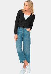 Schwarze Strickjacke, V-Ausschnitt, Knopfverschluss. Hellblaue, hochgeschnittene Jeans, cropped weit geschnitten. Weiße Sneaker. Insgesamt glatte Textur.