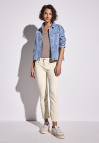 Giacca di jeans azzurro chiaro, adatta con bottoni e tasche, abbinata a pantaloni cropped beige e sneakers beige con dettagli bianchi.