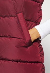 Ręka w kieszeni burgundowego pikowanego kamizelki puchowej noszonej na białej koszuli z długim rękawem.