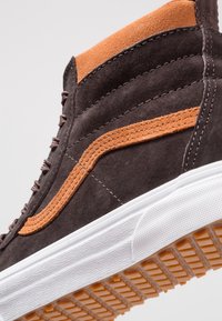High-Top-Sneaker aus dunkelbraunem Wildleder mit orangefarbenen Akzenten. Ausgestattet mit strukturierter weißer Gummisohle und kontrastierenden Steppnähten.