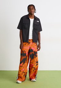 Sort kortærmet skjorte med to brystlommer, sat sammen med brede bukser i orange og sort med et abstrakt mønster, og hvide sneakers.
