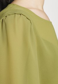 Grüne Bluse aus strukturiertem Stoff, mit leichten Raffungen an den Schultern und einer sanften, fließenden Silhouette. Schlichte, elegante Ausschnitt.