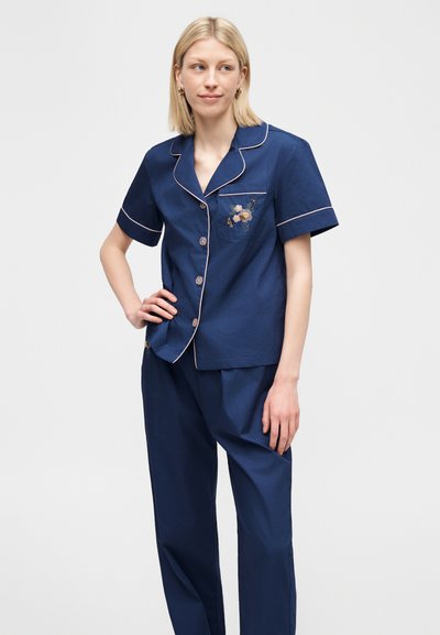 Γυναίκα με ξανθά μαλλιά που φοράει σετ πυτζάμας κοντομάνικο σε ναυτικό μπλε με απαλό ροζ περίγραμμα και κέντημα με λουλούδια στην τσέπη, στέκεται με το χέρι στη μέση.