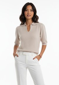 Beżowy sweter typu pulower z krótkim rękawem, z ostro zakończonym kołnierzem i dekoltem w kształcie klucza. Teksturowana tkanina i subtelne detale na rękawach. Białe spodnie.