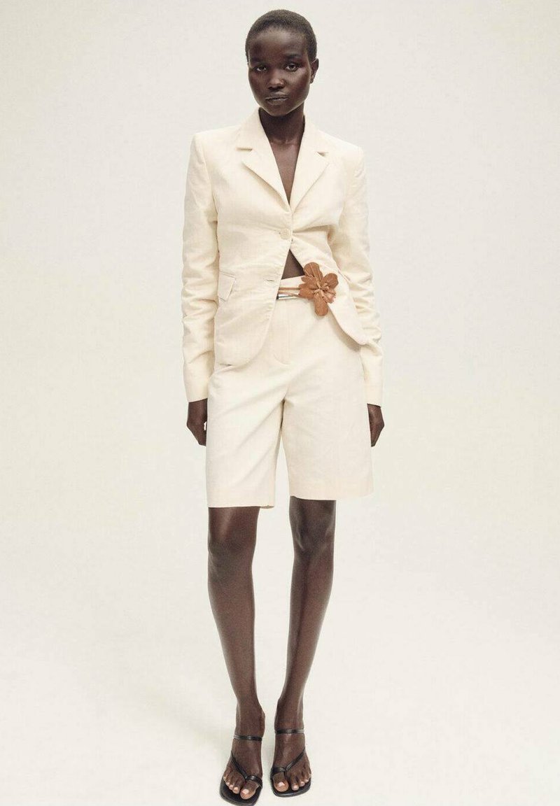 Beiger Leinen-Blazer mit cropped Schnitt, passenden Bermuda-Shorts und einem dekorativen braunen Blumen-Gürtel. Sandalen mit dünnen schwarzen Riemen.