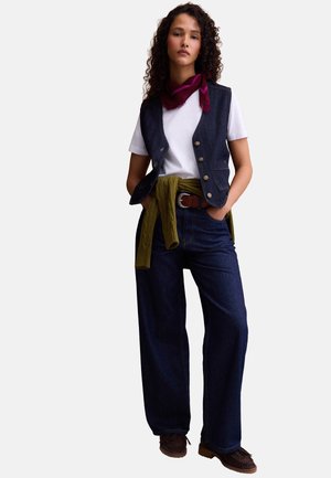 Donna che indossa una t-shirt bianca, un gilet blu scuro e jeans a gamba larga con un maglione verde legato in vita e mocassini marroni.