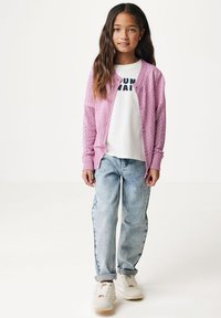 Ljusrosa stickad cardigan med ett spetsmönster över en vit t-shirt med svart text, kombinerad med ljusblå baggy jeans och vita sneakers.