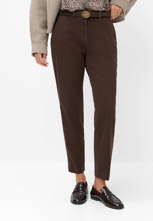 Person iført mørkebrune bukser med brun bælte, mønstret skjorte, beige jakke og sorte læder loafers på hvid baggrund.