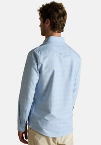 Muž oblečený v svetlomodrej, dlhými rukávmi, kockovanej košeli, zobrazený zozadu, skombinovanej s béžovými nohavicami na jednoduchom pozadí.