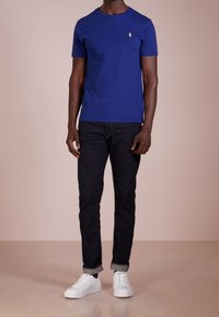 Královsky modré tričko s krátkým rukávem vyrobené z bavlny, s kulatým výstřihem a malým zlatým logem na hrudi, spárované s tmavými džíny a bílými teniskami.