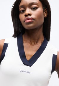 Jeune femme aux cheveux noirs et raides portant un haut blanc sans manches avec un col en V bleu marine et le logo "Calvin Klein" sur la poitrine.