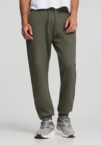 Oliwkowe joggery zwężane z miękkiego materiału z elastycznym pasem. Mankiety przy kostkach, zestawione z szarymi butami sportowymi.