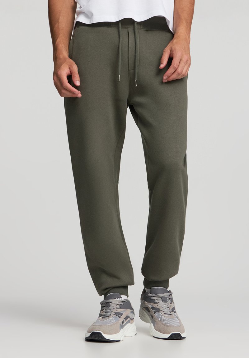 Oliwkowe joggery zwężane z miękkiego materiału z elastycznym pasem. Mankiety przy kostkach, zestawione z szarymi butami sportowymi.