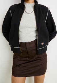 Černá zipová bunda s vysokým límcem, šedý svetr pod ní a hnědá sukně. Bunda má kontrastní švy a nášivku s logem.