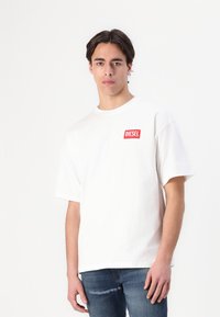 T-shirt en coton blanc avec un col rond, arborant un logo carré rouge avec le texte "DIESEL". Manches courtes et coupe décontractée. Porté avec un jean bleu.