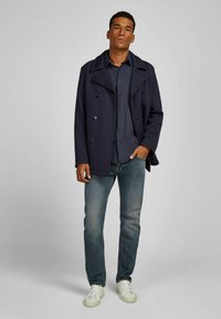 Marineblauem, doppeltknöpfigem Mantel mit großen Knöpfen, über einem dunklen Hemd. Hellblaue Jeans und weiße Sneaker runden das Outfit ab.