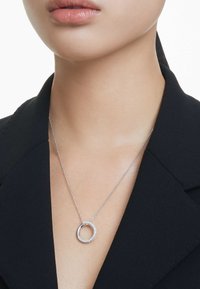 Collar de plata con un diseño circular, que presenta un borde texturizado y una banda interna lisa. Lleva una cadena de plata contra una blusa oscura.