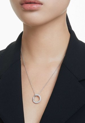 Collana con pendente in argento dal design circolare, caratterizzata da un bordo texturizzato e da una banda interna liscia. Indossata su una catena argentata contro un top scuro.