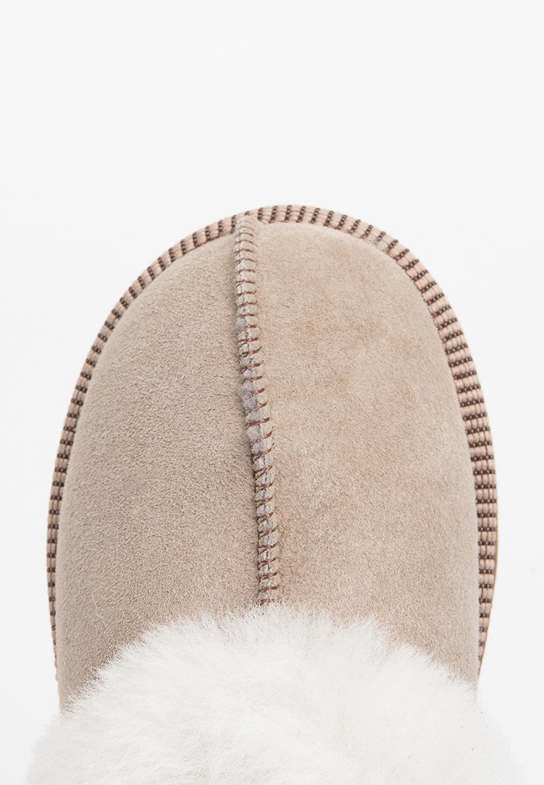 Pantofola in suede beige con punta arrotondata, dotata di un morbido rivestimento bianco e cuciture a contrasto lungo il perimetro. Suola piatta.