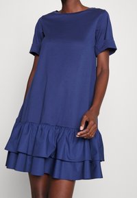 Marinblå bomullsklänning med rundad halsringning och korta uppvikta ärmar. Har en flätad volangnederkant för extra volym och textur.
