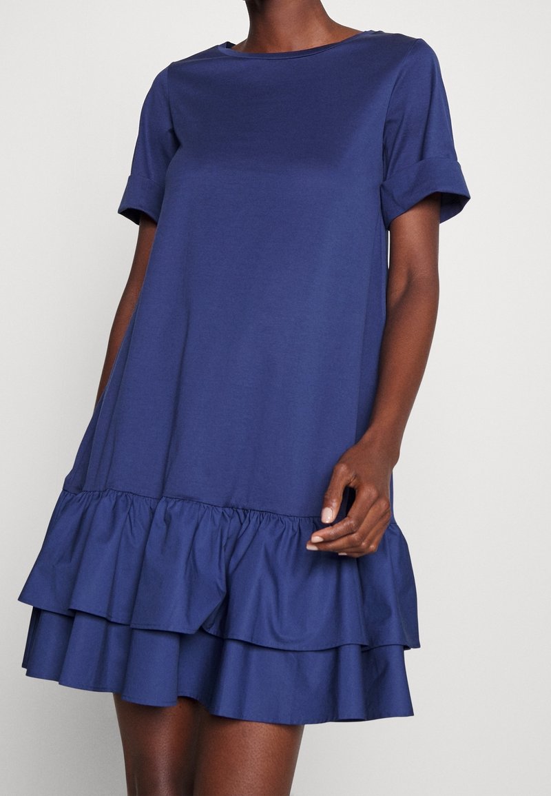 Marinblå bomullsklänning med rundad halsringning och korta uppvikta ärmar. Har en flätad volangnederkant för extra volym och textur.