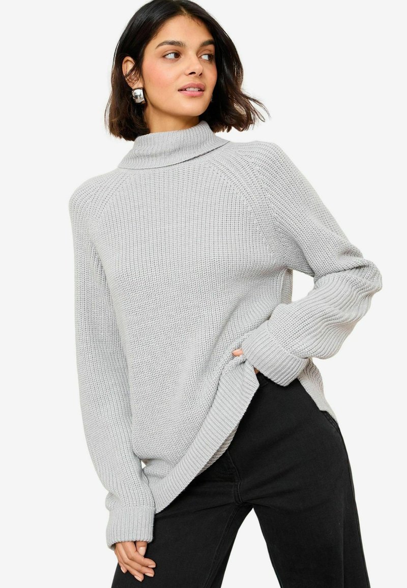 Hellgrauer Strickpullover mit hohem Kragen, gerippten Bündchen und seitlichen Schlitzen. Über schwarzen Jeans getragen. Strukturiertes Maschenmuster sichtbar.