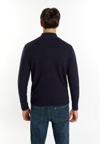 Temno moder pleteni pulover z visokim ovratnikom, rebrastimi manšeti in robom, v kombinaciji s srednje opranimi jeans hlačami. Udobna tekstura, prilegajoč dizajn.