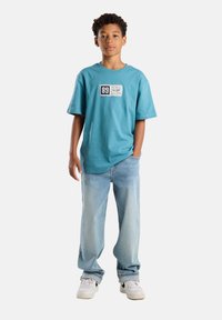 Turkoosi puuvillapaita, jossa on suorakulmainen paikka, jossa on merkintä "89". Yhdistetty haalistuneisiin sinisiin farkkuihin ja valkoisiin lenkkareihin, joissa on mustia yksityiskohtia.